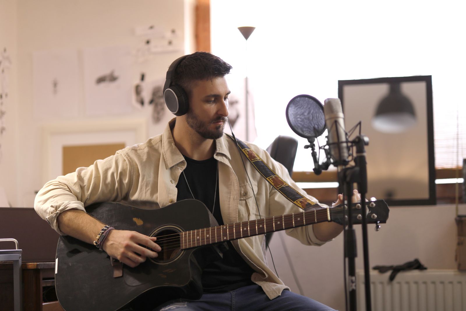 A musician in headphones playing acoustic guitar into a microphone at home