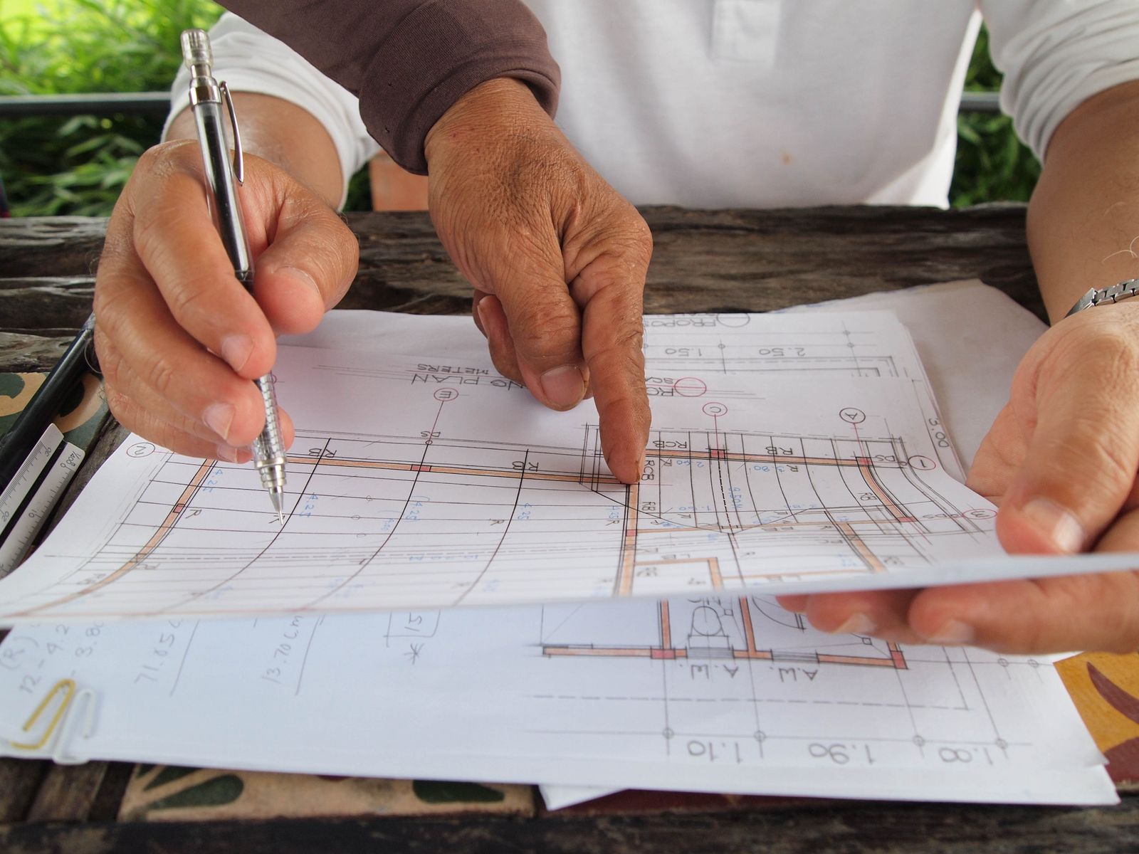 Two people going over printed house plans with a pencil on a work table