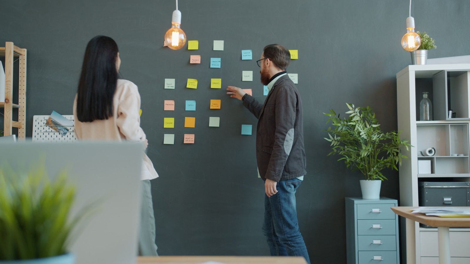 Two colleagues arranging sticky notes on a dark wall in an office