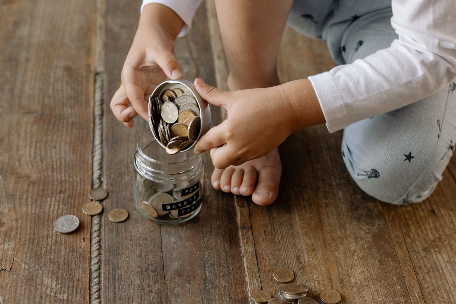 A child's hands putting coins into a glass jar on a wooden floor