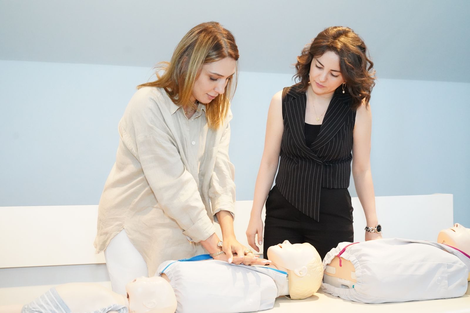 A trainer watching a learner practise CPR on a manikin, with more manikins on the table