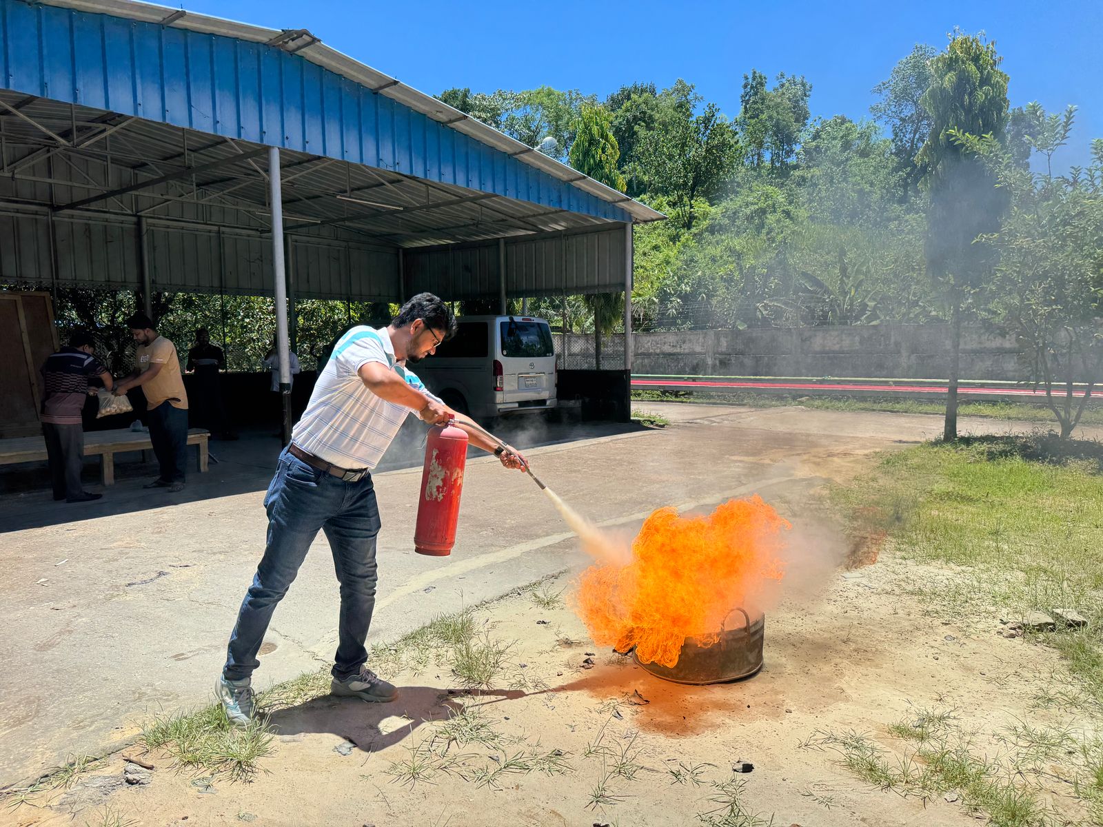A man aiming a red fire extinguisher at a controlled fire in a metal pan during outdoor practice