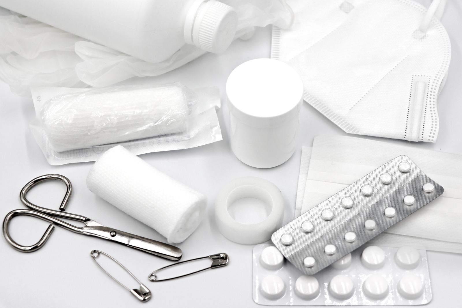 First-aid supplies laid out on a white table: bandages, tape, scissors, safety pins, a mask and blister packs