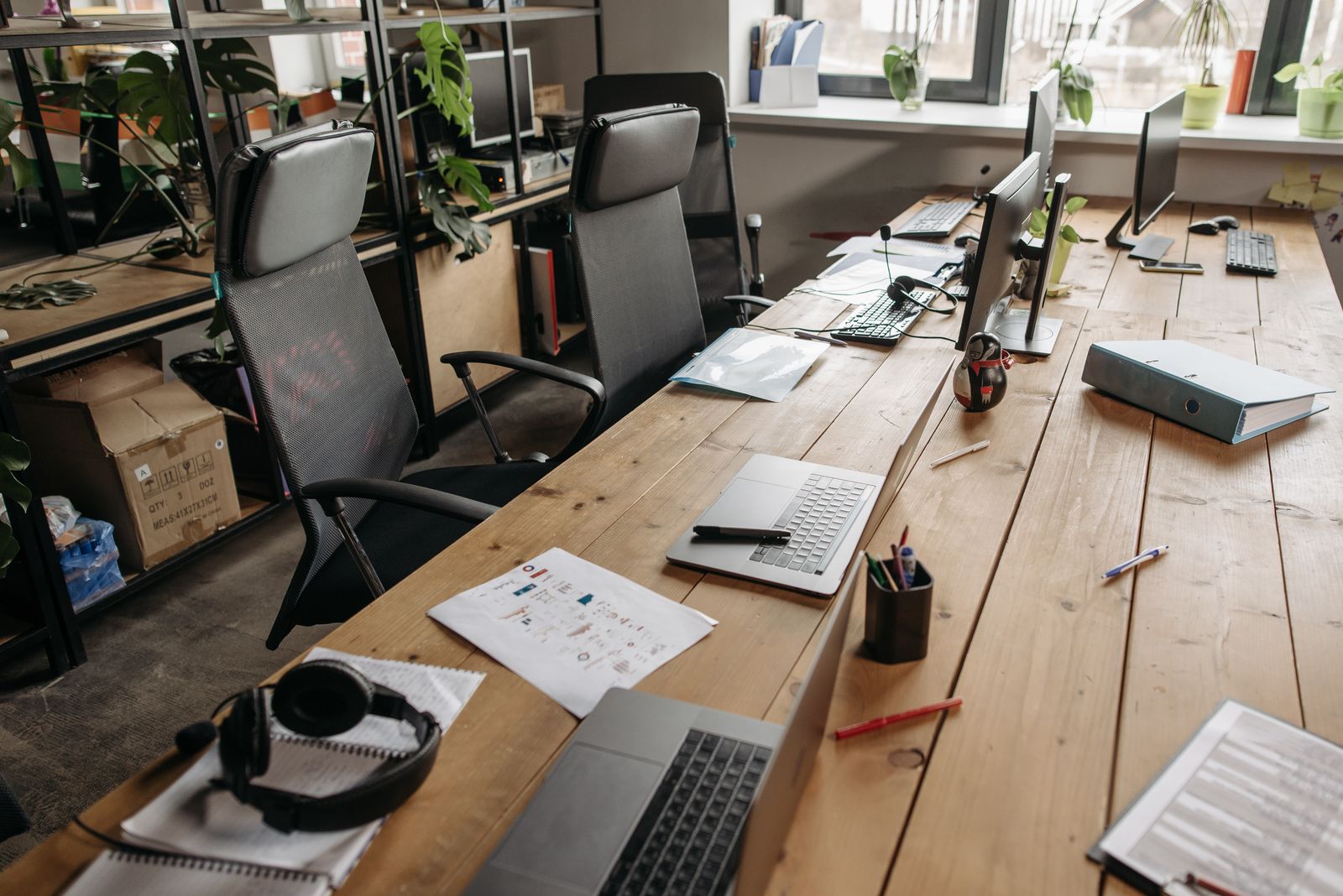 A long wooden bench of shared desks with laptops, chairs and monitors in a bright coworking room