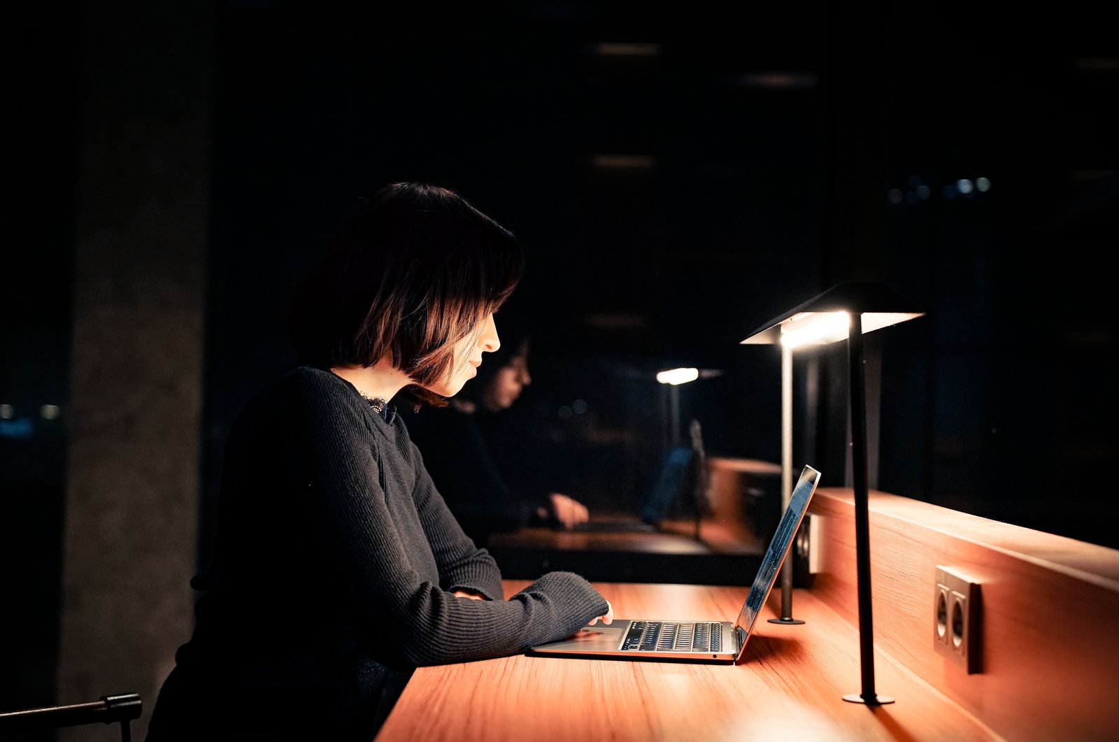 An engineer working at a laptop at a desk lit by one lamp late at night