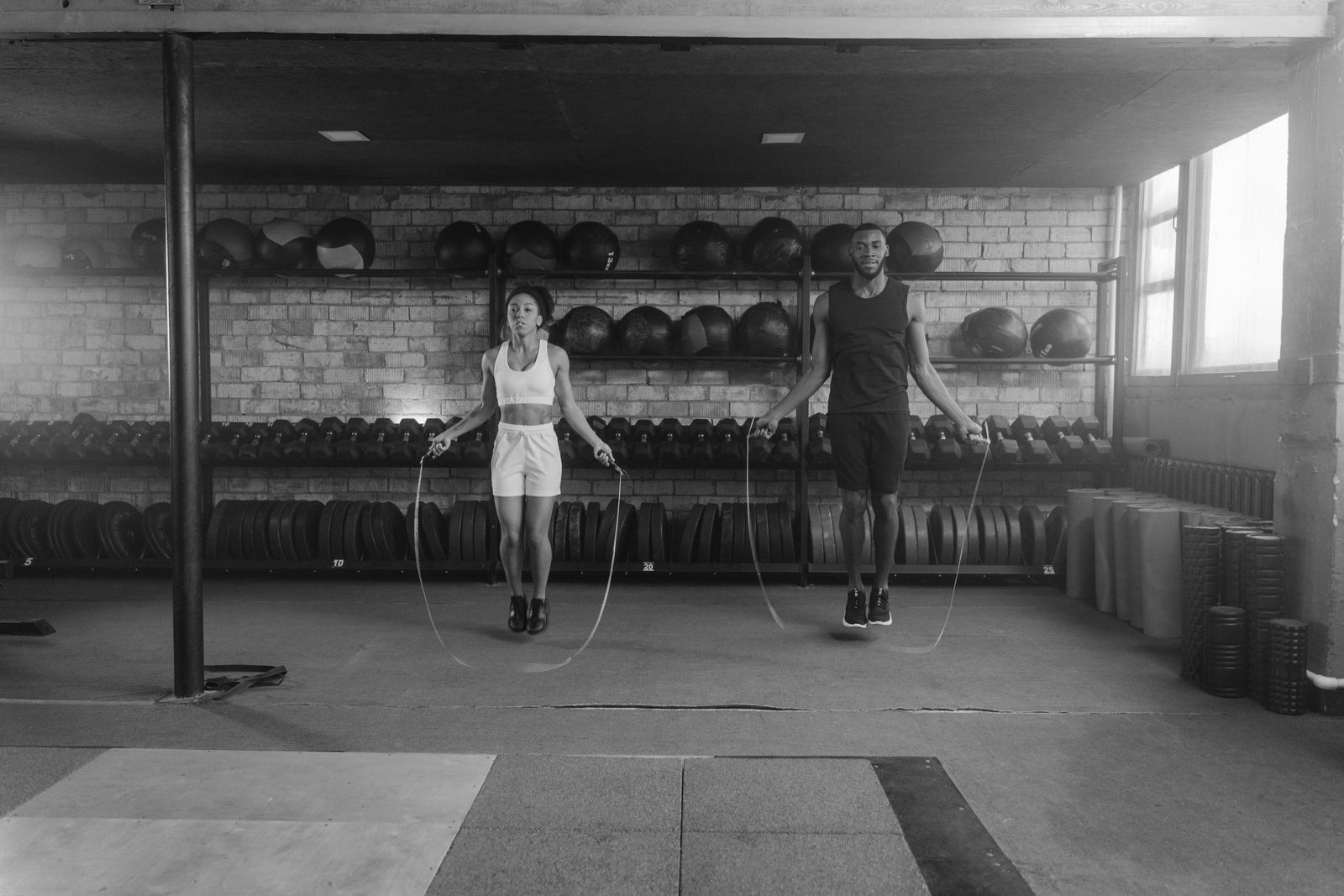 Black and white photo of two people skipping in a gym with medicine balls on the shelves behind