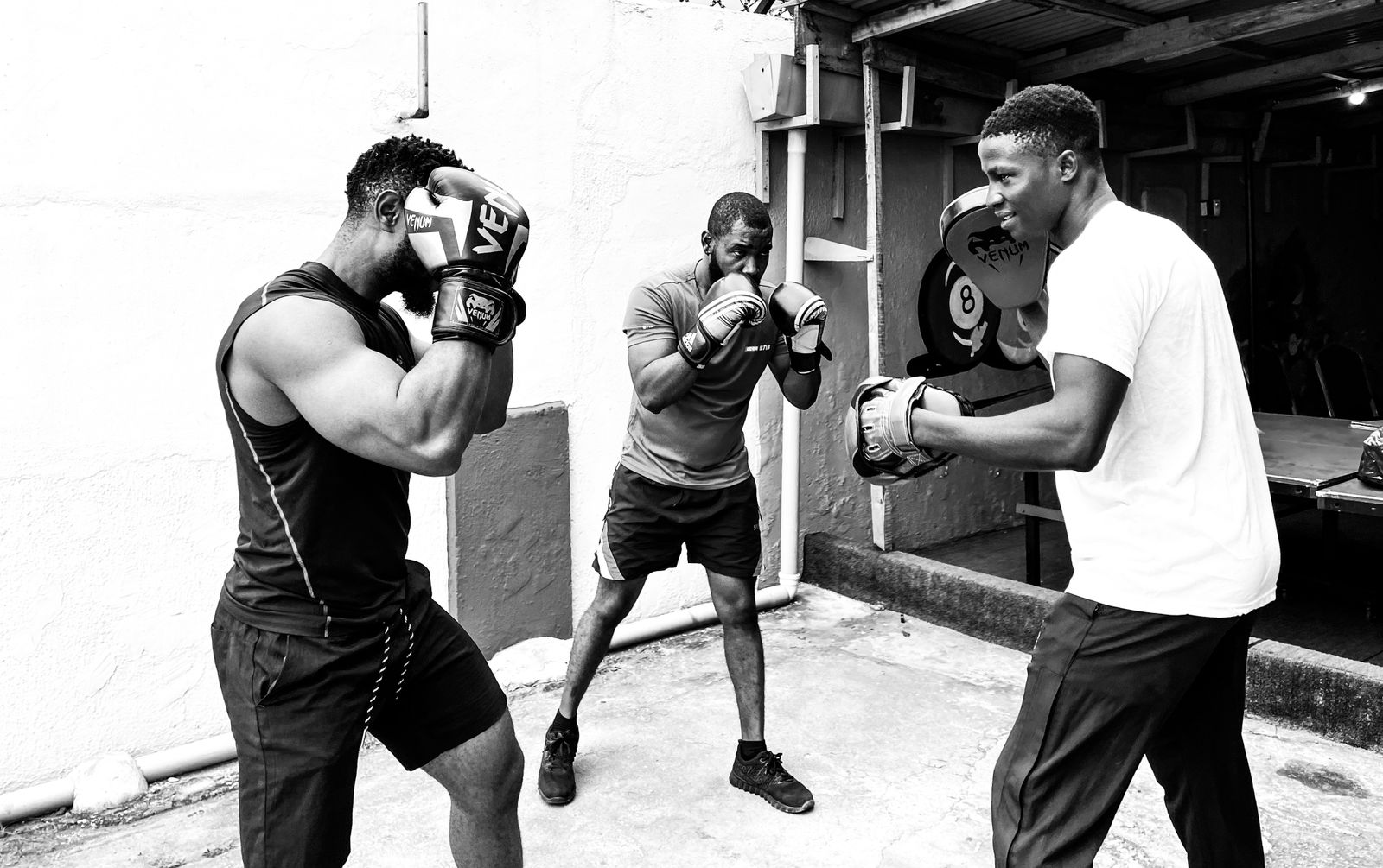 Black and white photo of a coach holding pads for a boxer while a third boxer shadowboxes behind