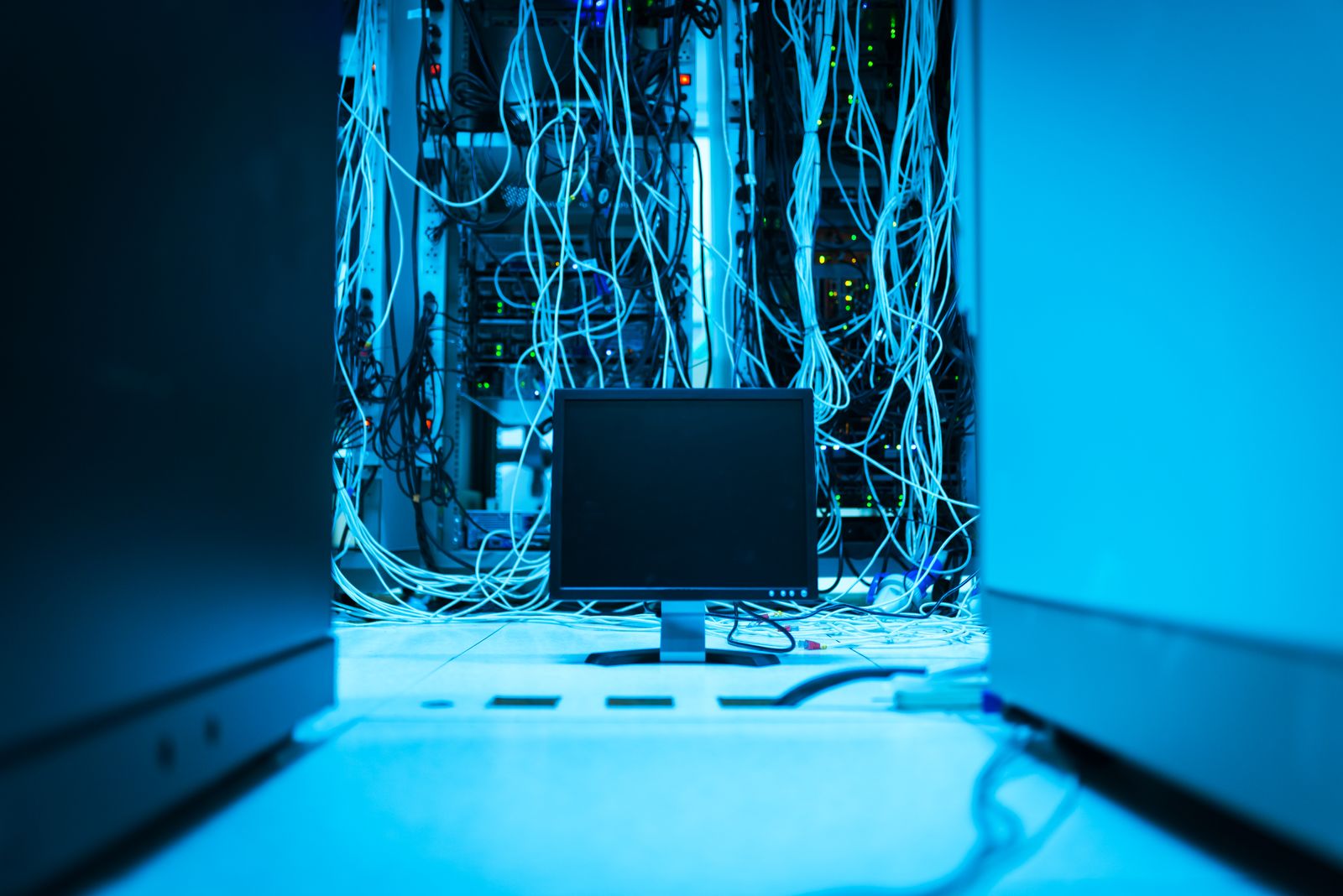 Bundles of network cables hanging behind a monitor in a blue-lit server room