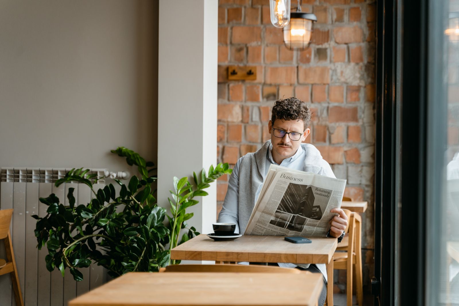 A man reading a newspaper at a café table by a brick wall