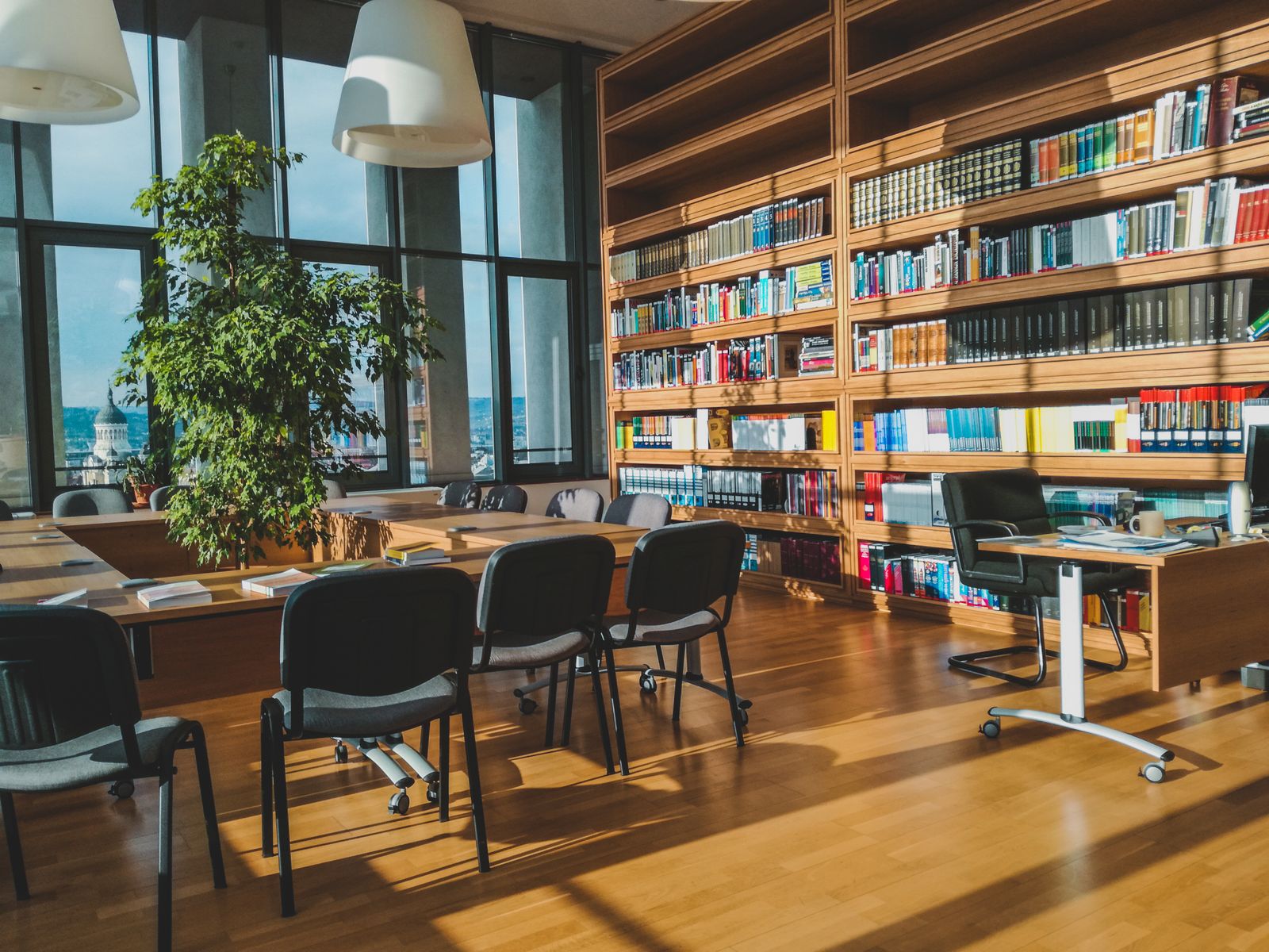 A sunlit library reading room with long tables, chairs and tall bookshelves