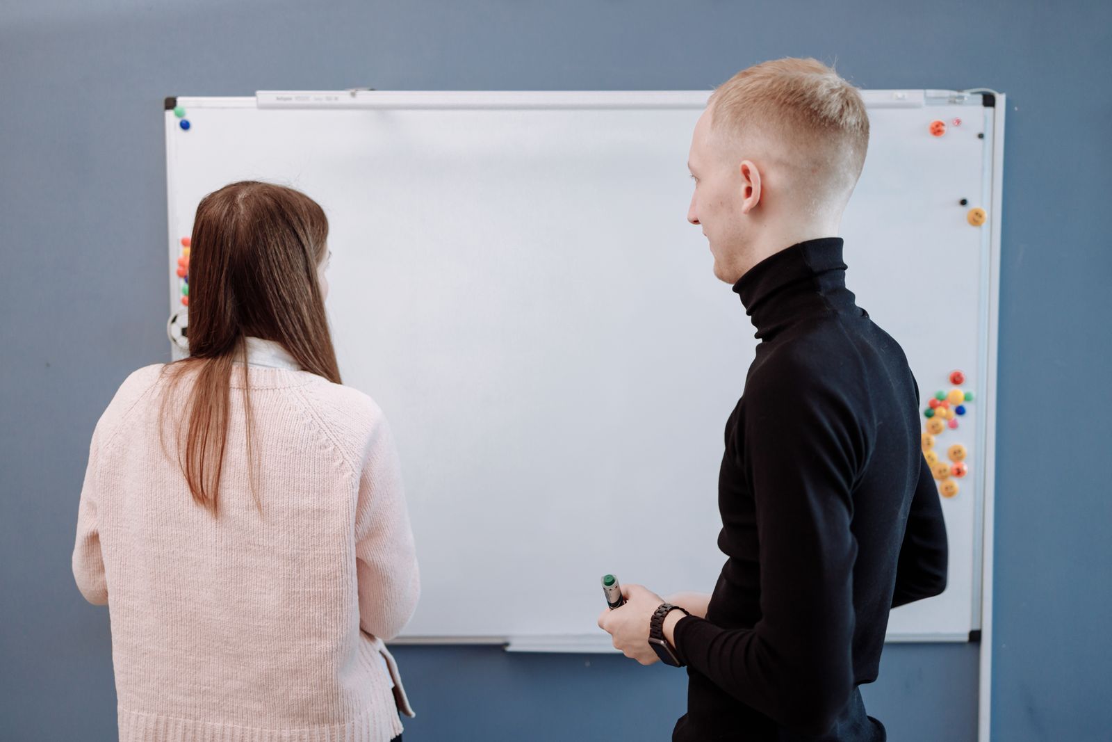 Two people talking in front of a whiteboard in a meeting room, seen from behind