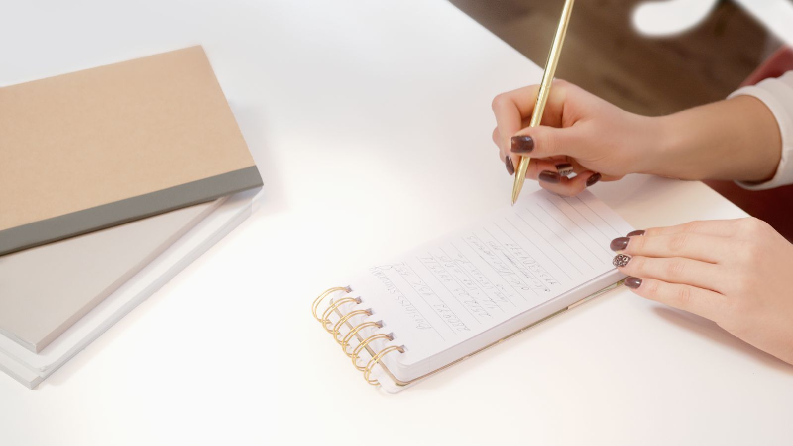 A hand writing with a pencil in a small spiral notebook on a white desk