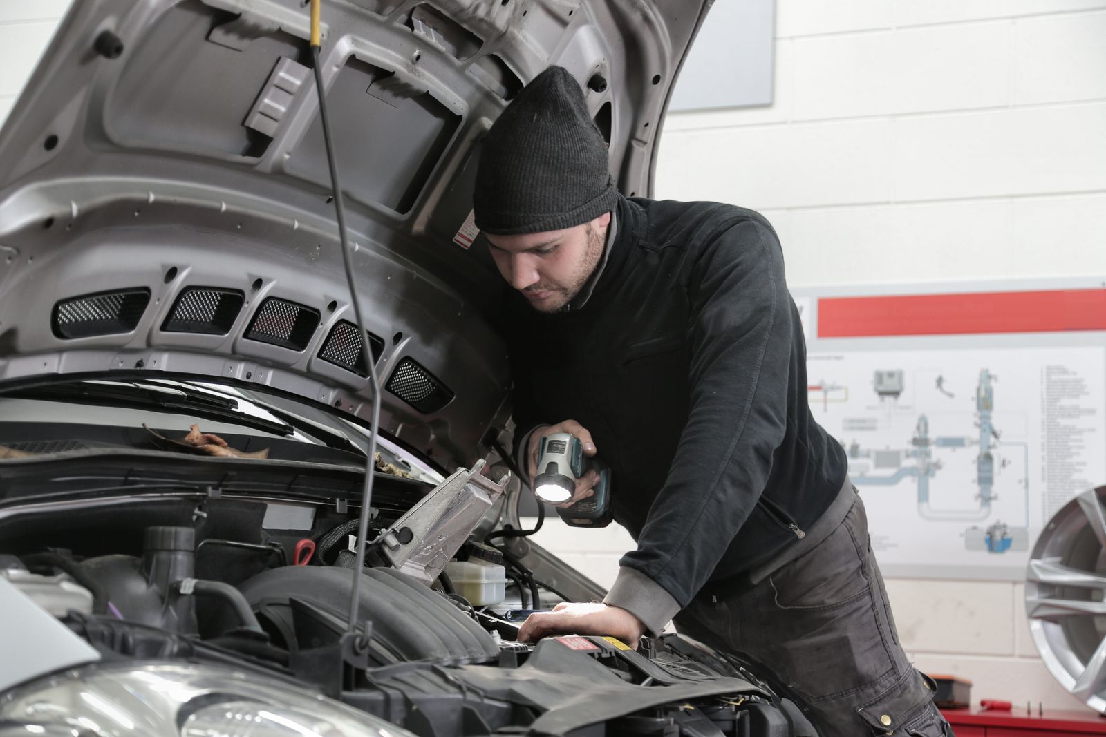 A mechanic in a knit cap leaning into an open engine bay with a flashlight