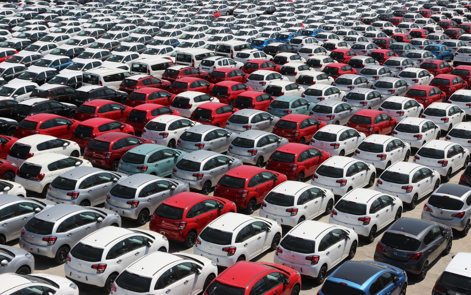 A car lot seen from above, rows of parked cars in white, grey, red and blue