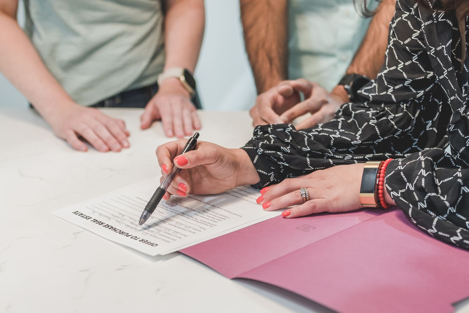 Hands signing a printed document on a white table beside a pink folder