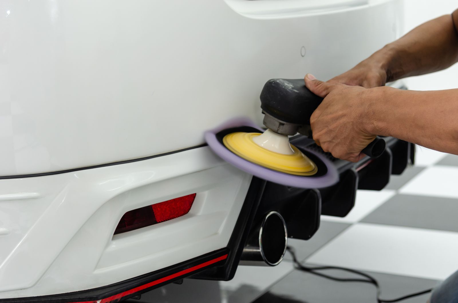 A detailer polishing the rear bumper of a white car with a power polisher