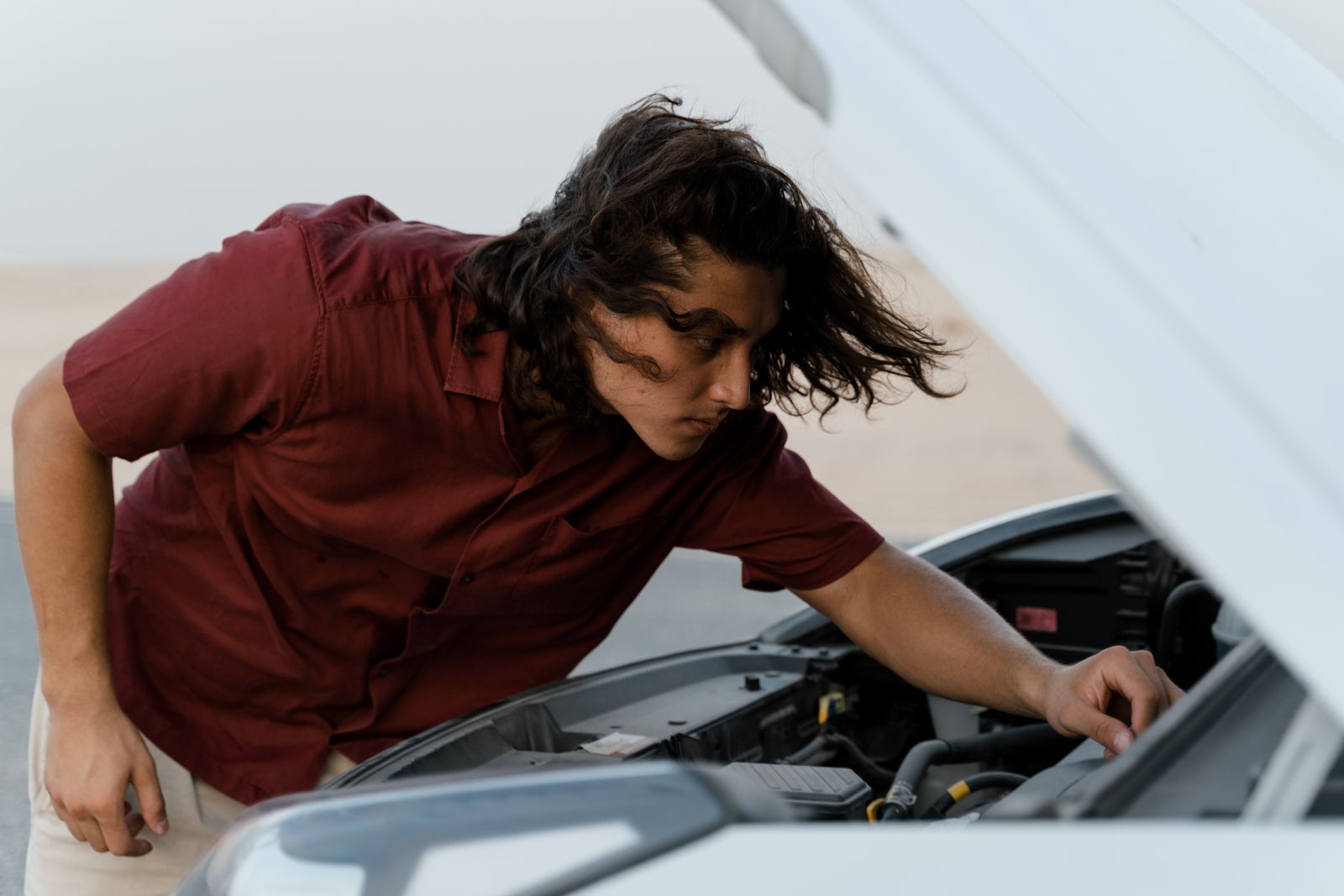 A person checking under the open hood of a white car outdoors on a bright day