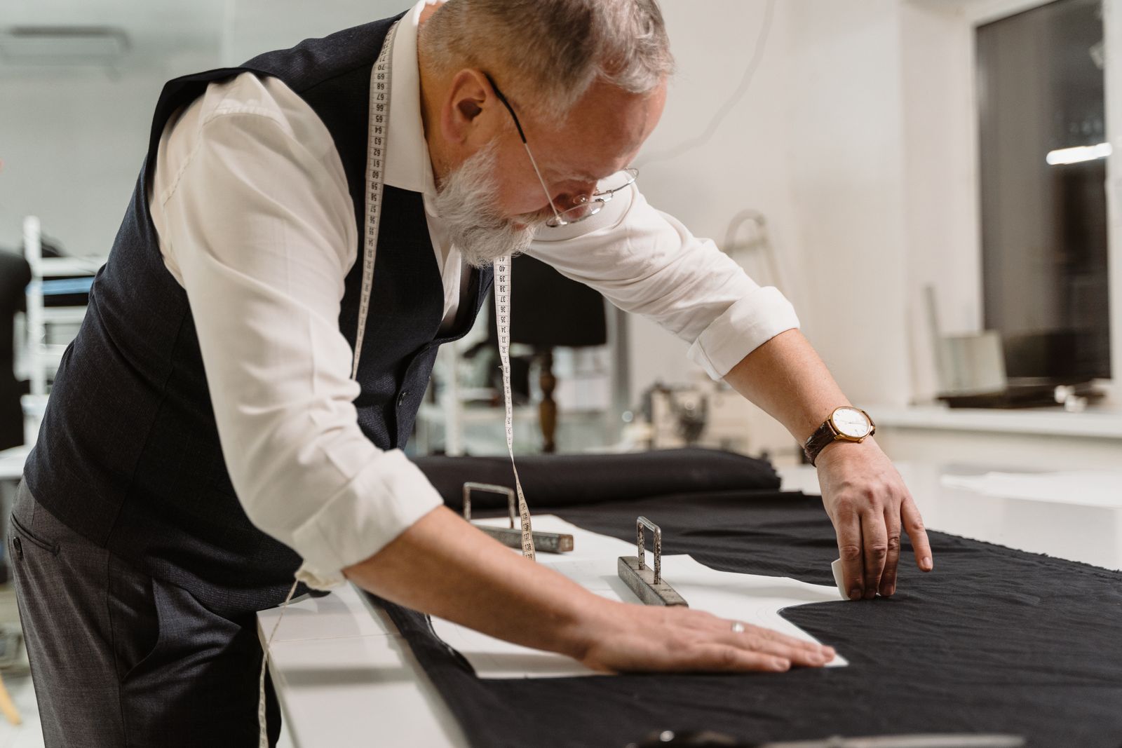 A grey-haired tailor in a waistcoat marking dark cloth on a cutting table