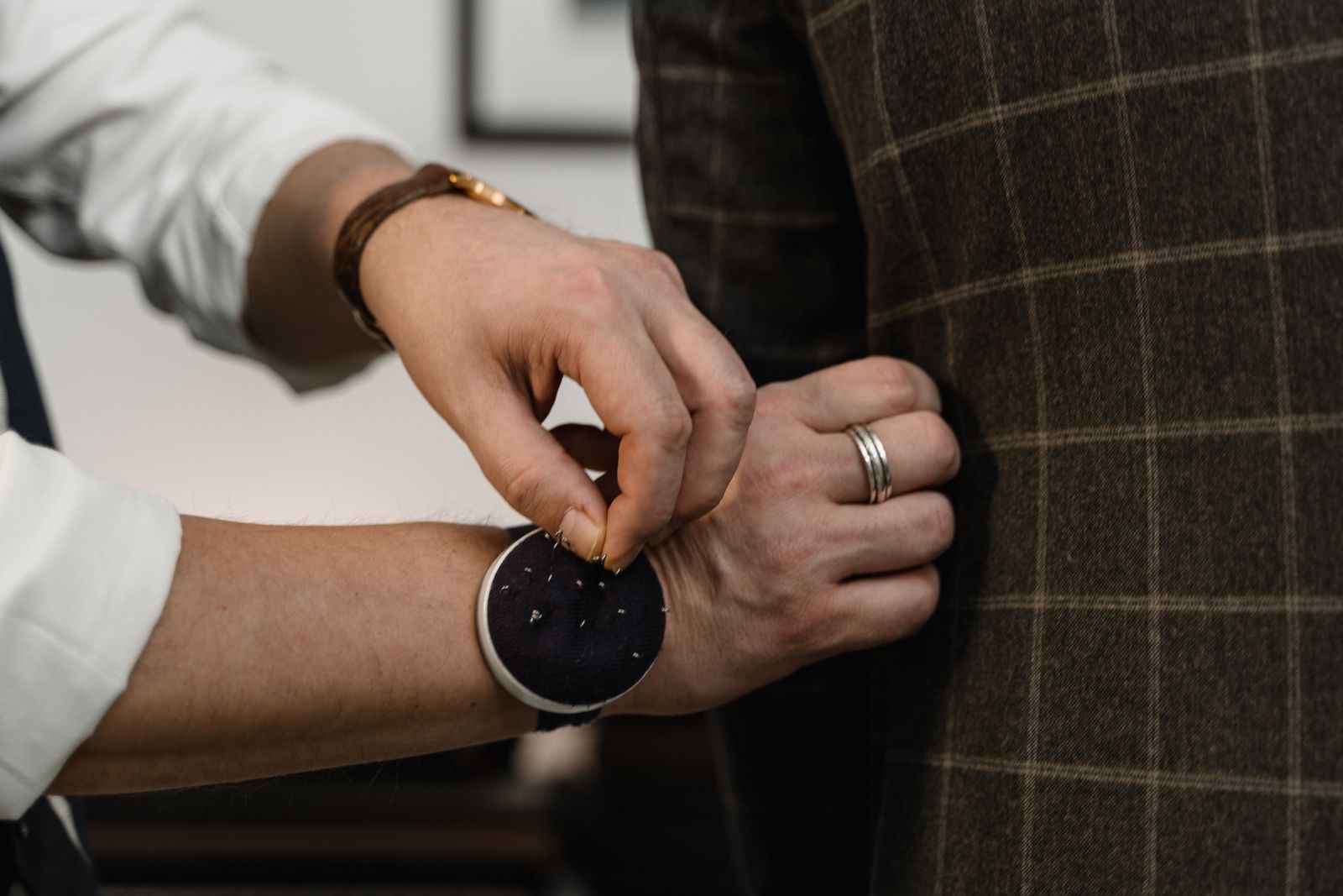 A tailor's hands taking a pin from a wrist pincushion to pin a brown check jacket