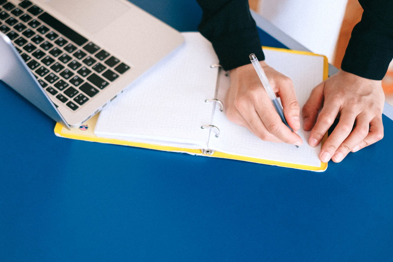 Hands writing in a notebook next to a laptop on a blue table