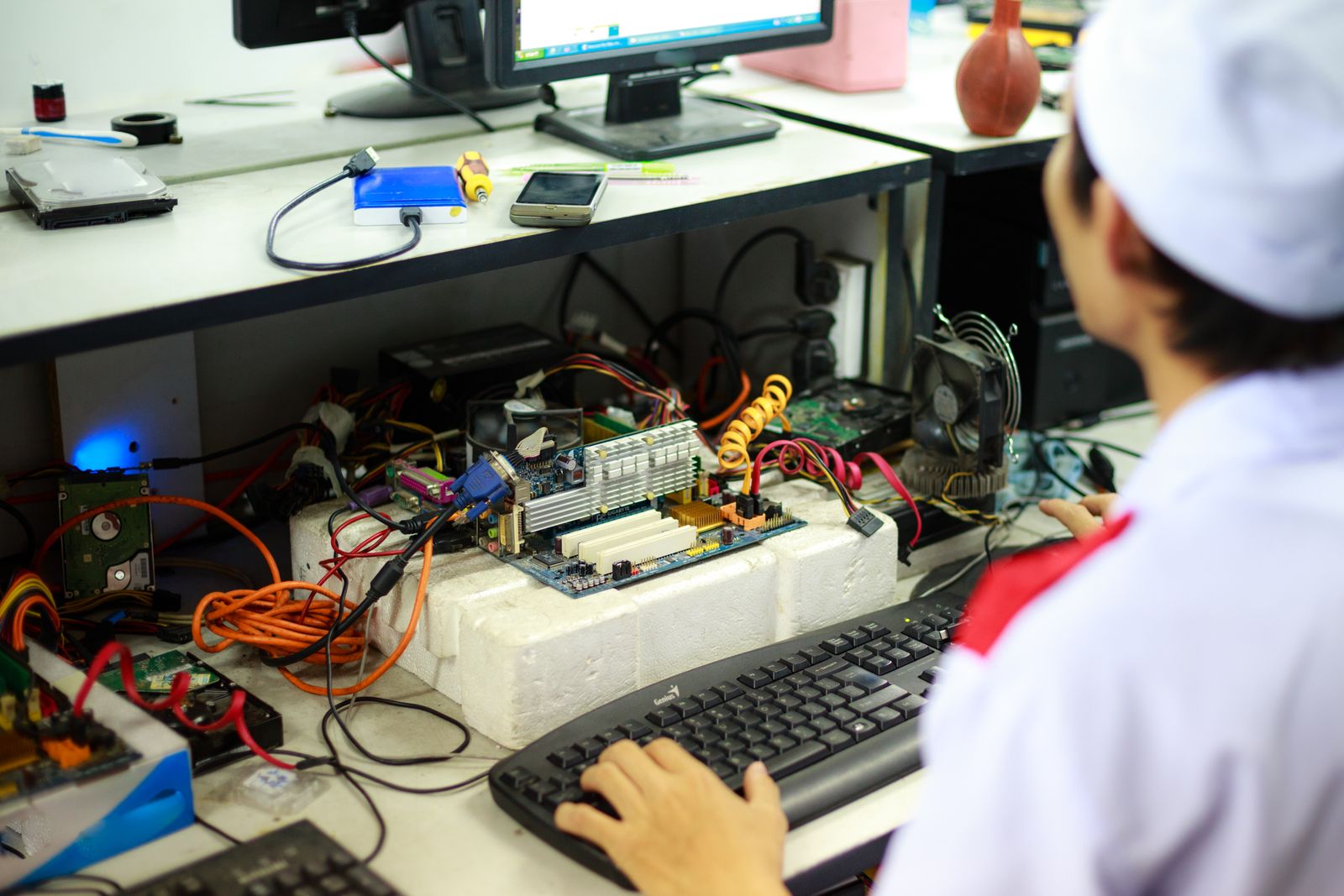A technician at a workstation full of boards, cables and test gear, seen from behind