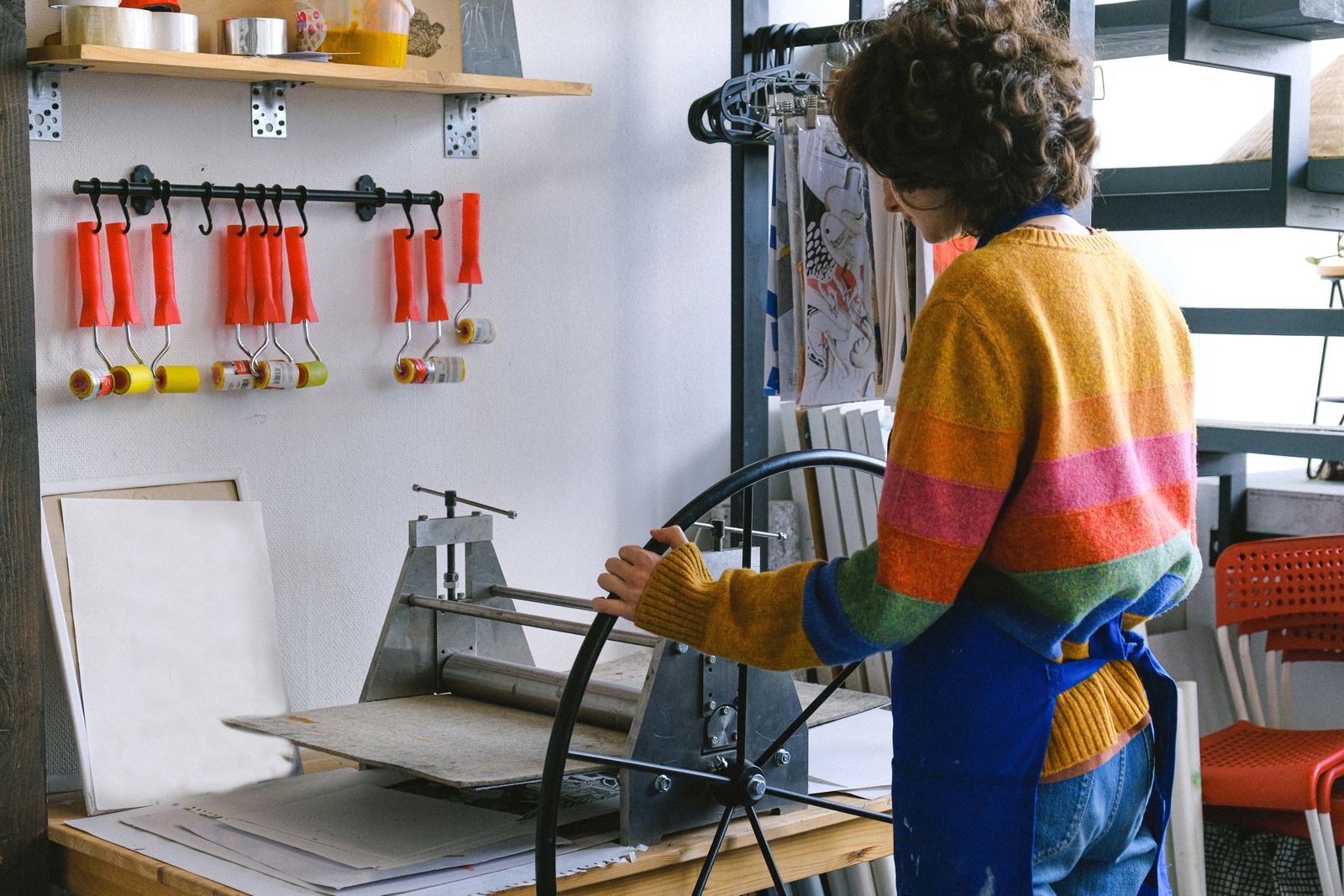 A printmaker in an apron turning the wheel of a print press in a bright studio