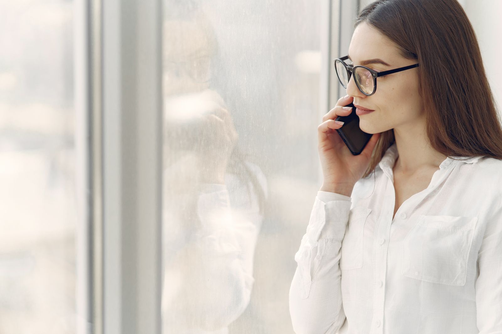 A woman in glasses talking on her phone by a bright window at home