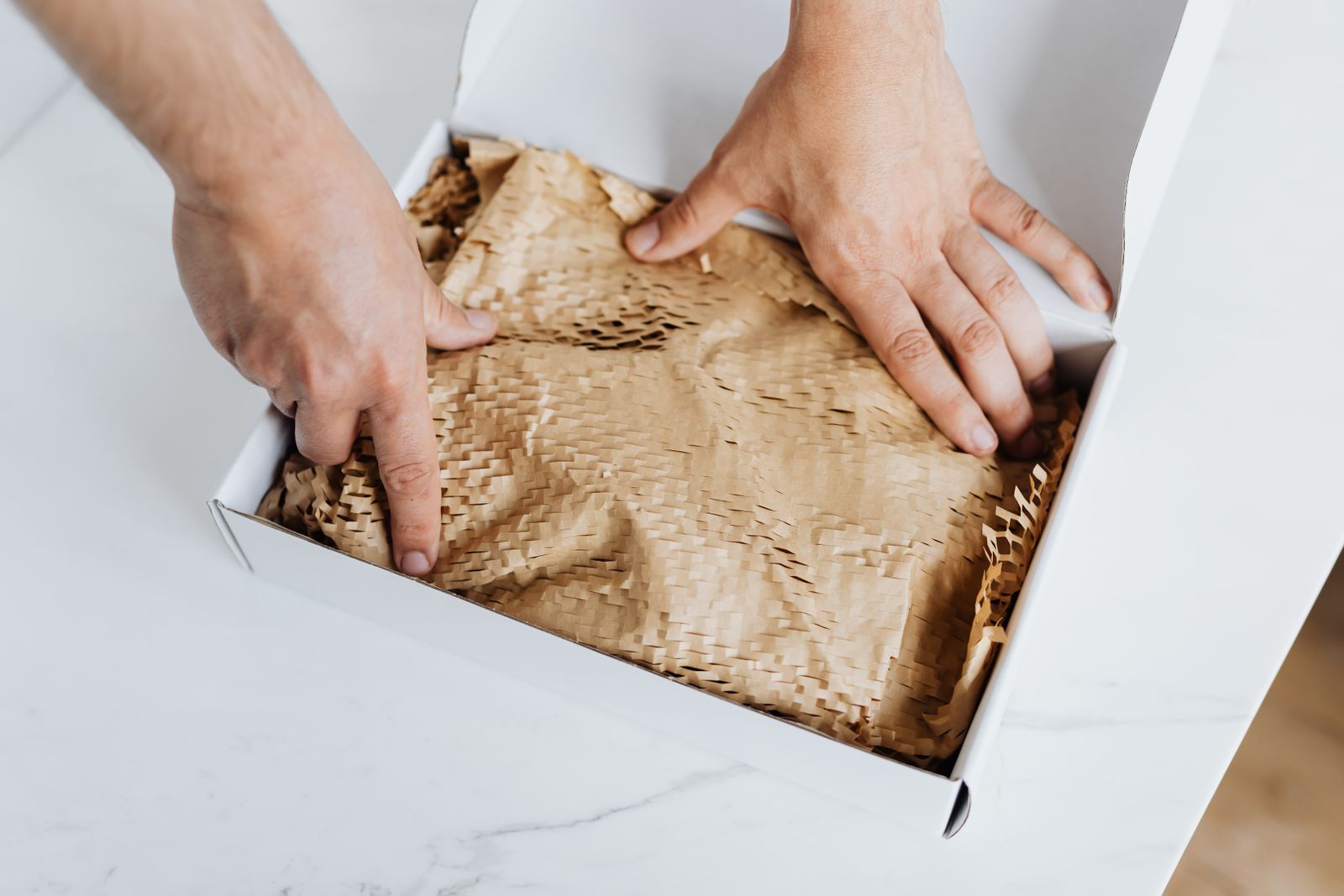 Hands packing a white box with brown crinkled paper on a marble table