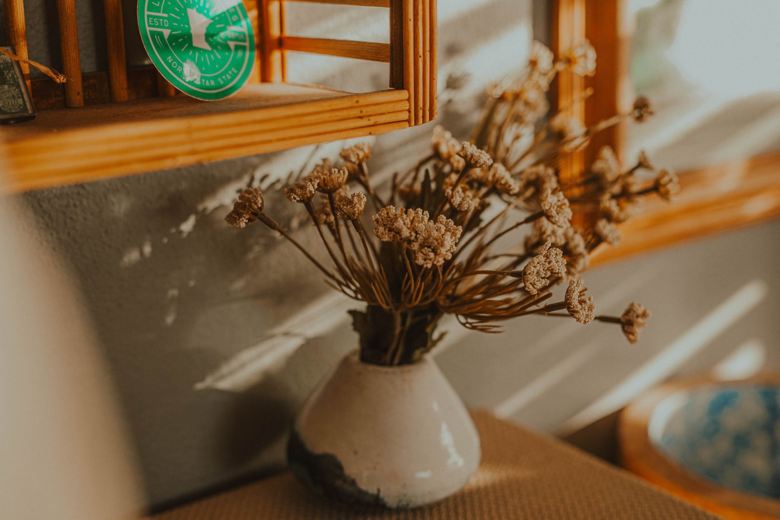 A white vase of dried flowers on a wooden shelf in afternoon sun