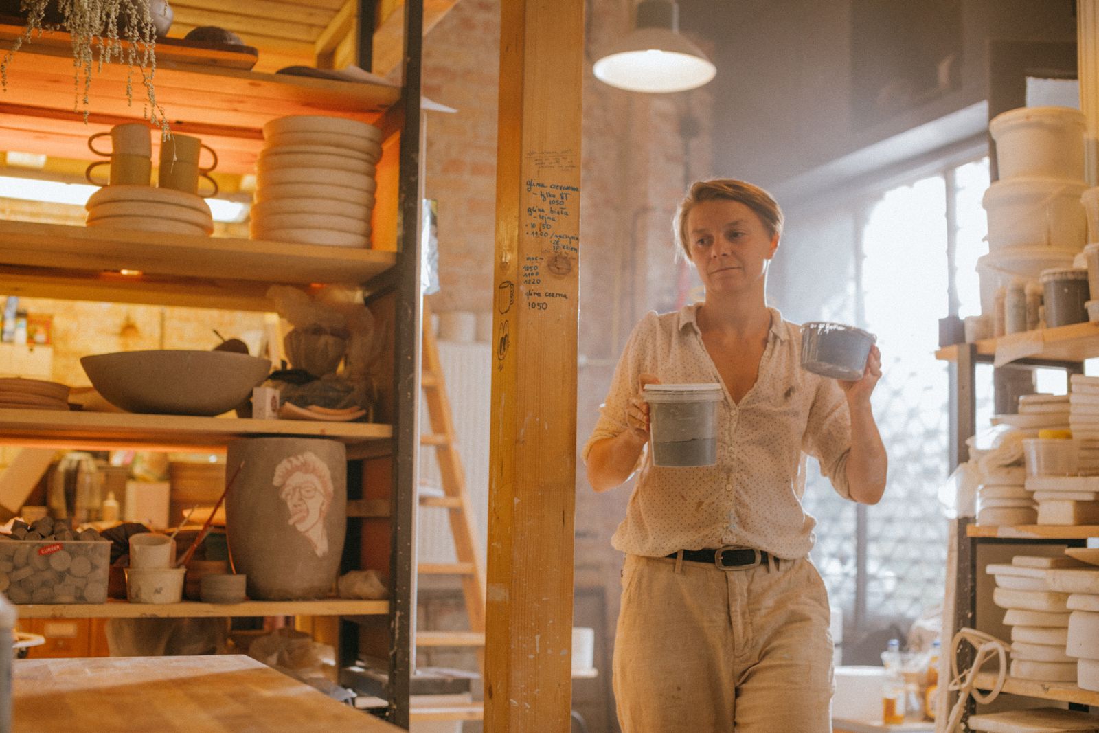 A potter holding two glazed bowls in a sunlit workshop with full shelves behind her