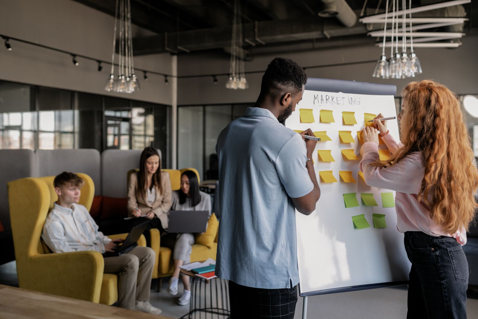 A small team planning at a whiteboard covered in notes while others work on laptops