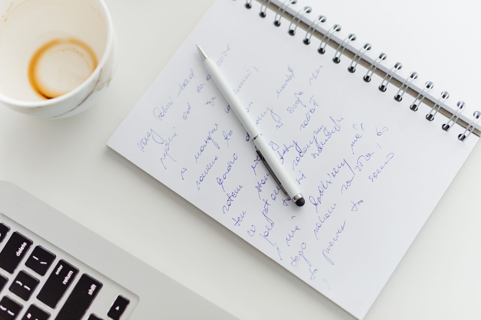 A notebook of handwritten notes and a pen beside a laptop and a coffee