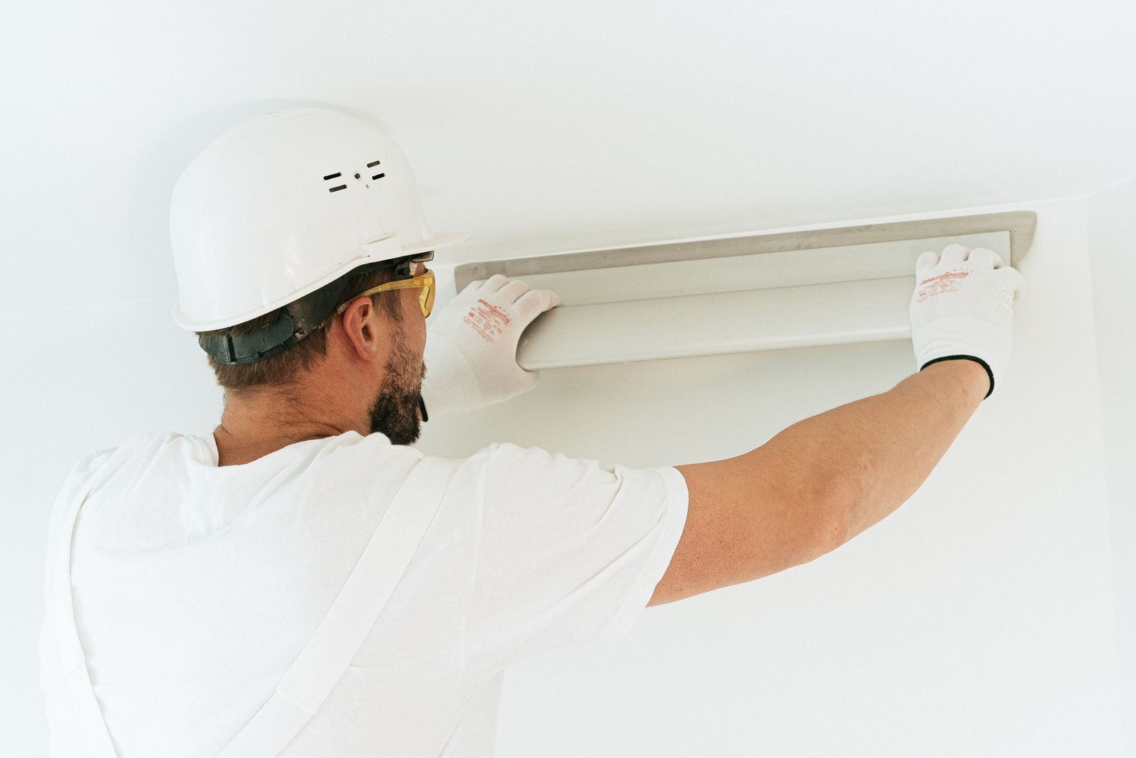 An electrician in a white hard hat fitting a light fixture high on a wall