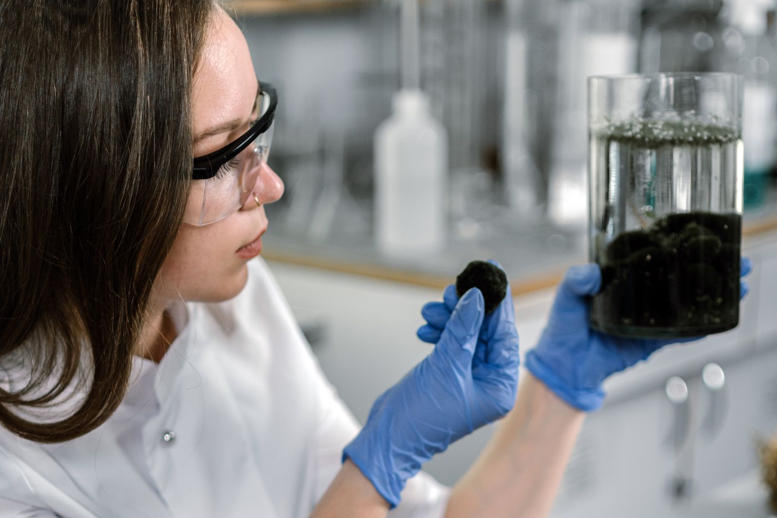 A lab analyst in gloves holding up a jar of soil and a soil sample