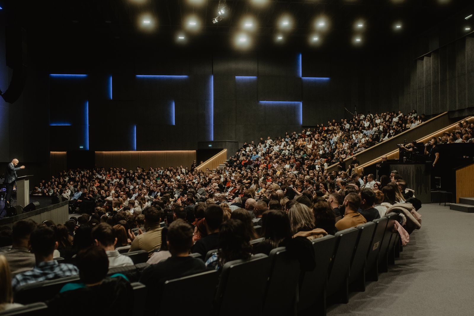 A full conference hall seen from the back rows, the audience facing a lit stage and screens