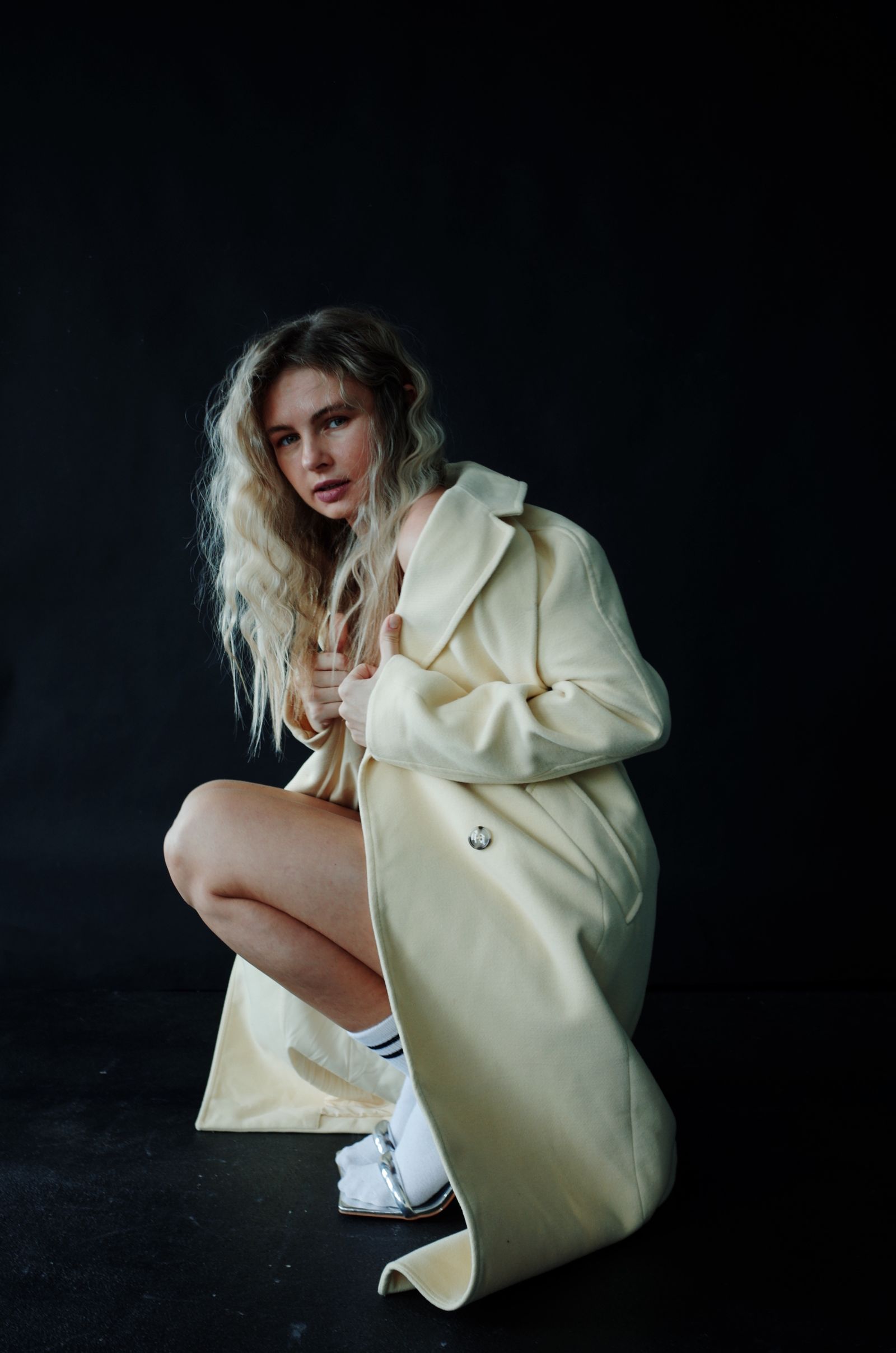 A model in a cream double-breasted wool coat crouching against a black backdrop