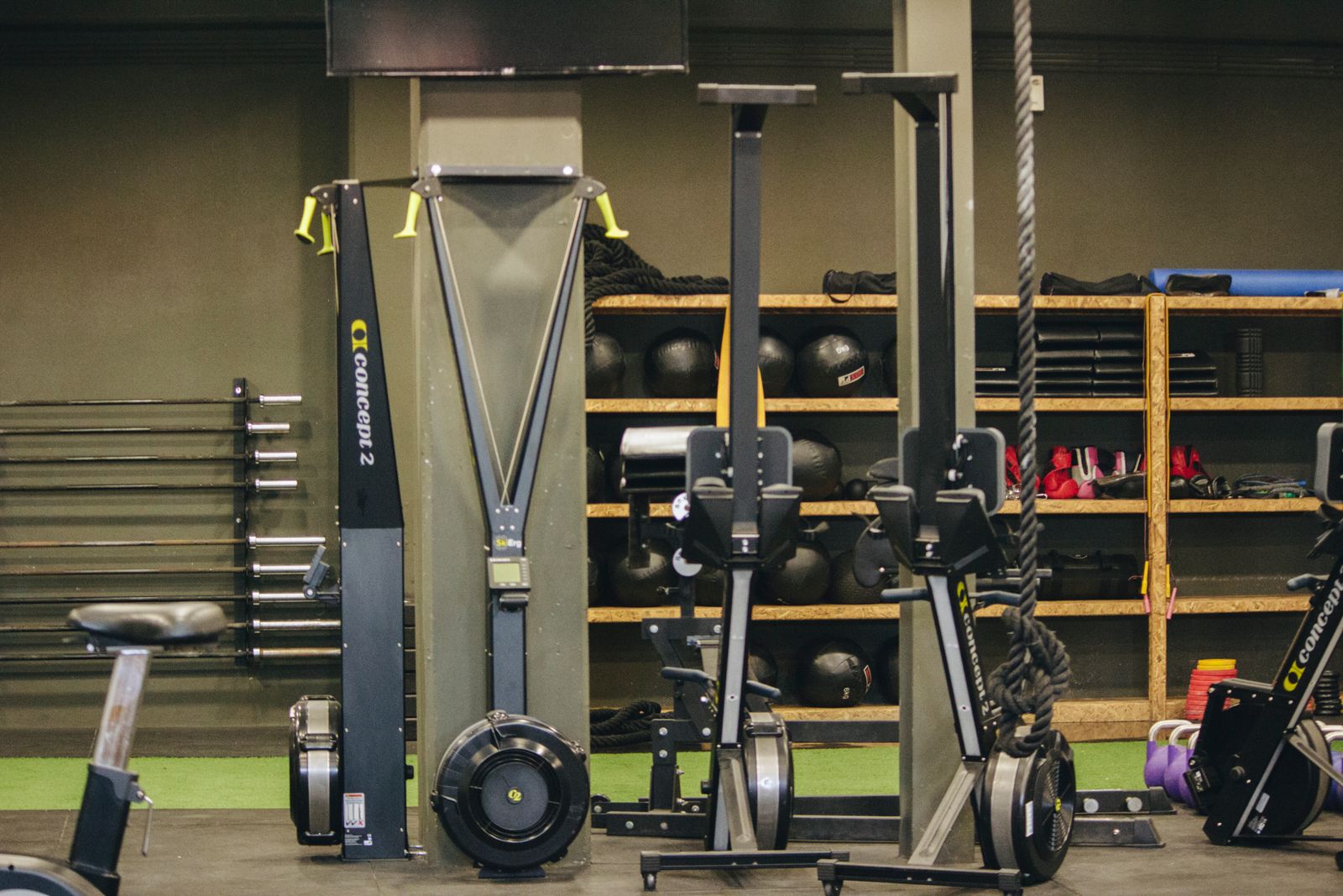 Gym floor with rowing machines, ski ergs, a rig and wall shelves of medicine balls