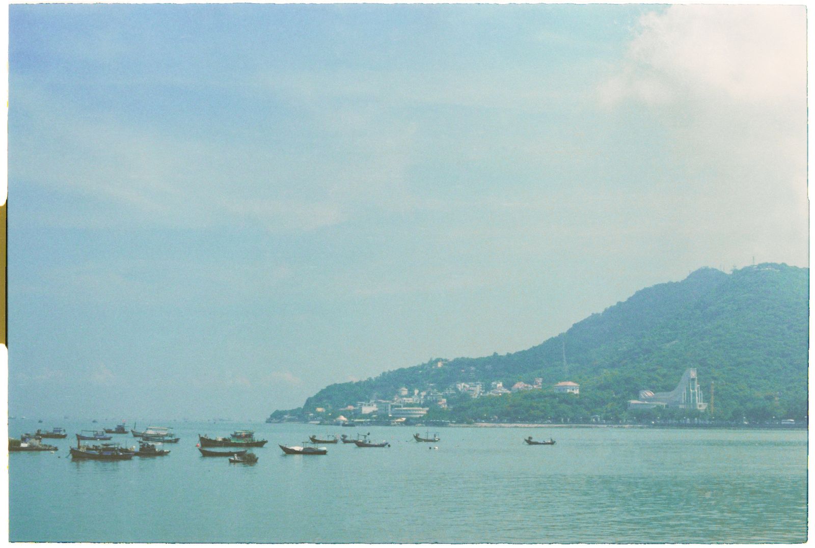A hazy bay with small boats and green hills, in pale washed-out colour