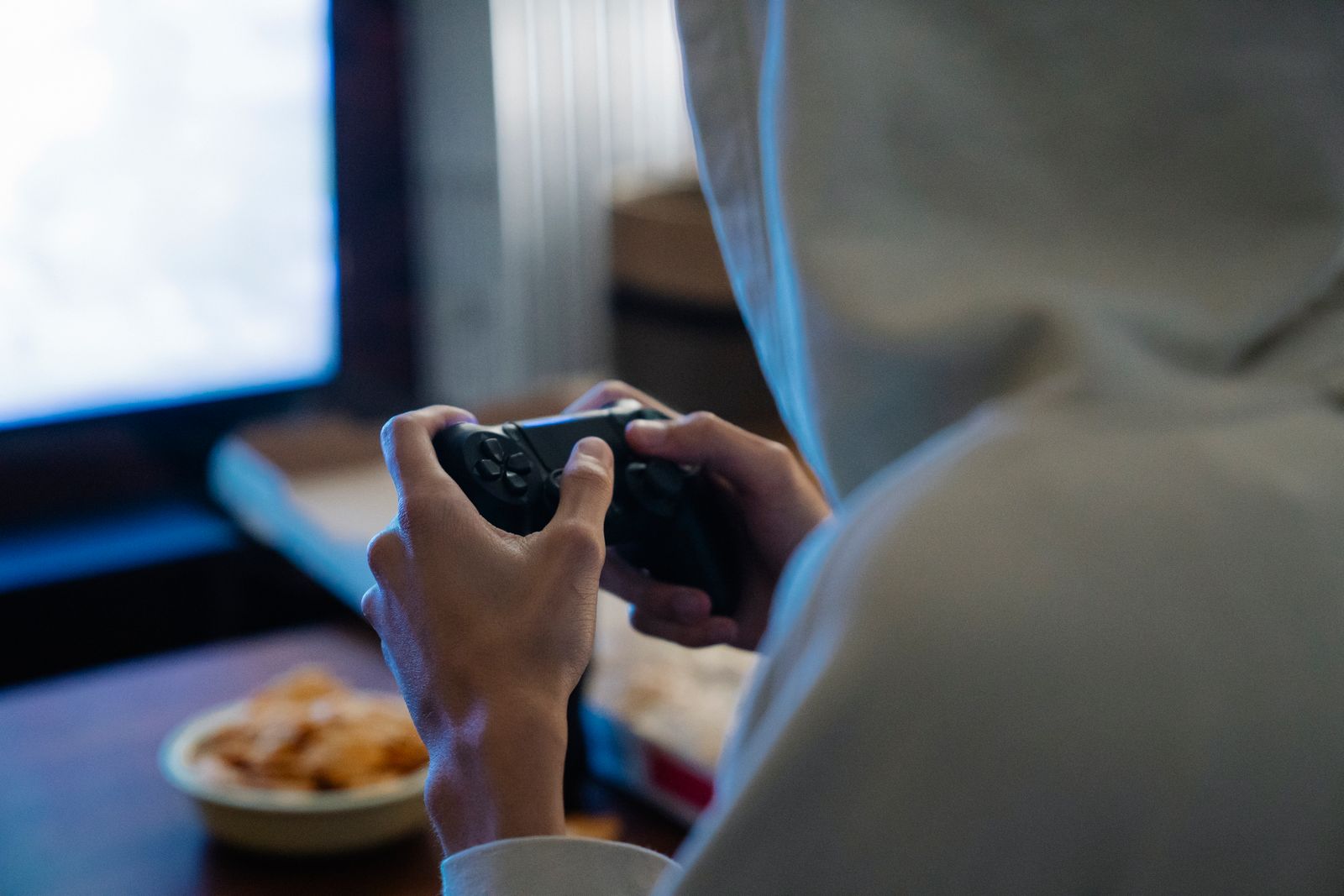 A player in a grey hoodie, seen from behind, playing with a gamepad in front of a monitor
