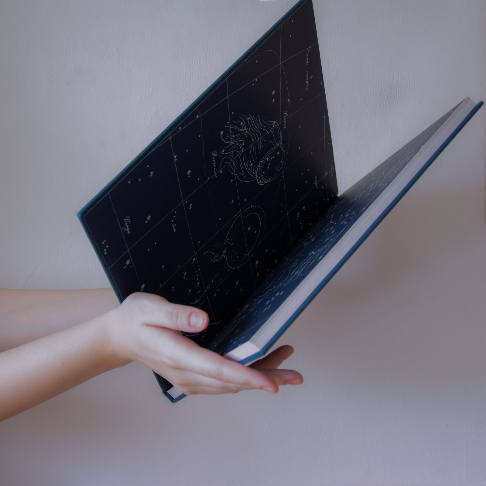 A hand holding open a star atlas with constellations drawn on dark blue pages