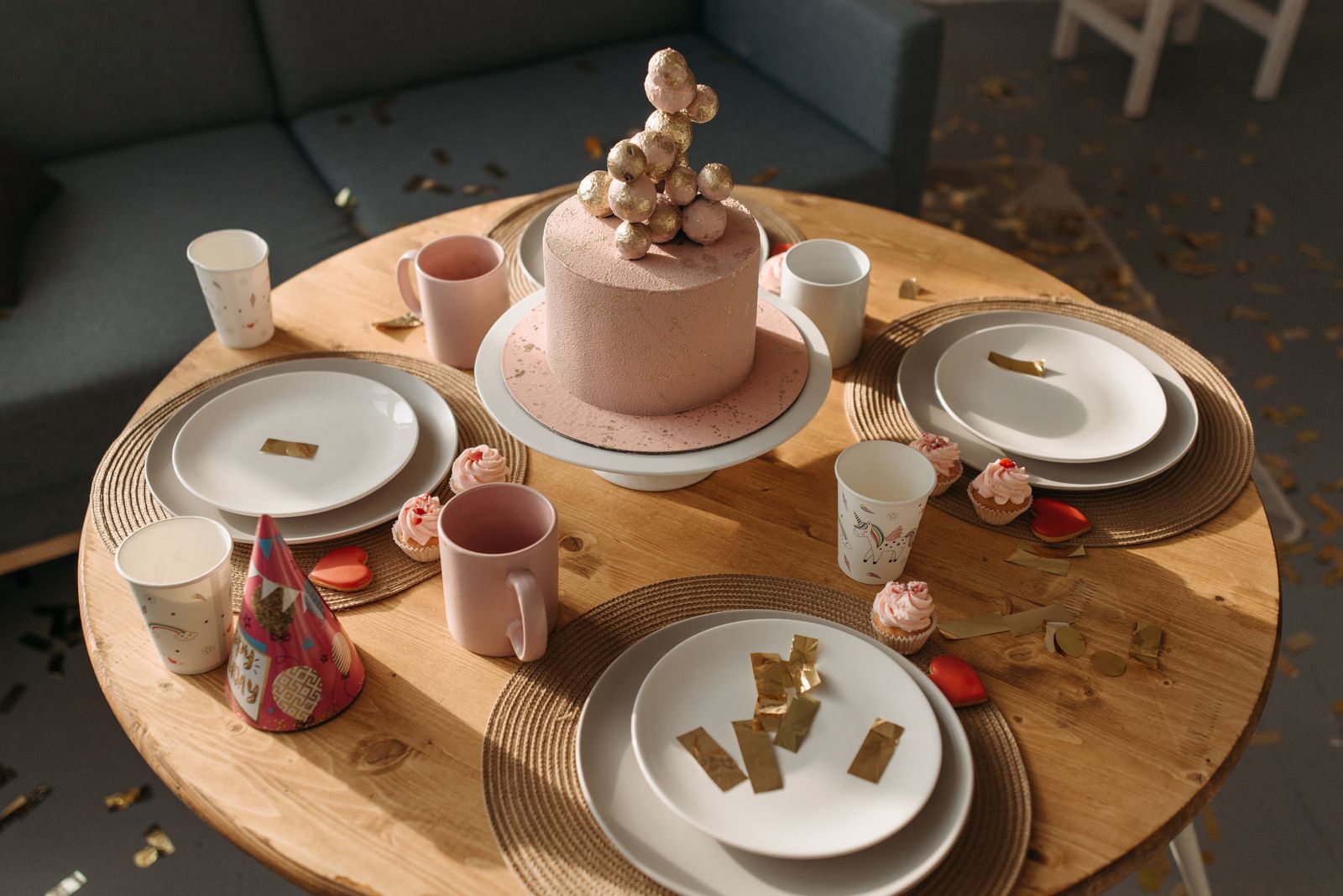 A party table with paper plates, cups, a pink cake and party hats on a wooden top