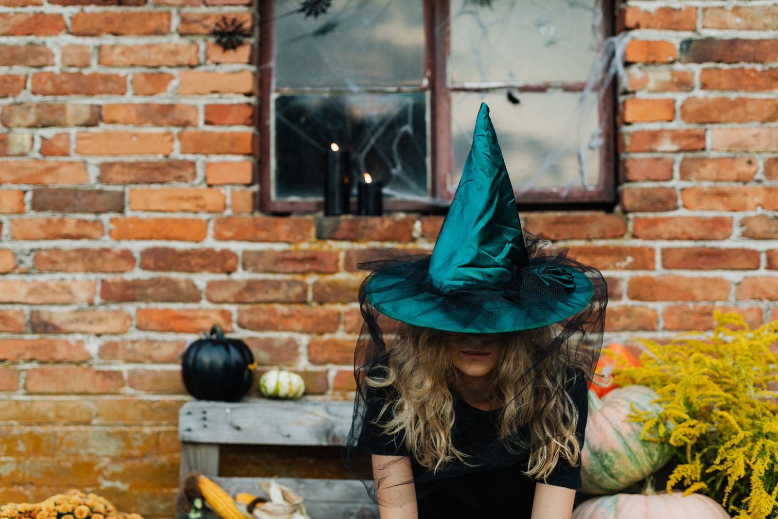 A woman in a teal witch's hat sitting against a brick wall beside small pumpkins, her face hidden by the brim