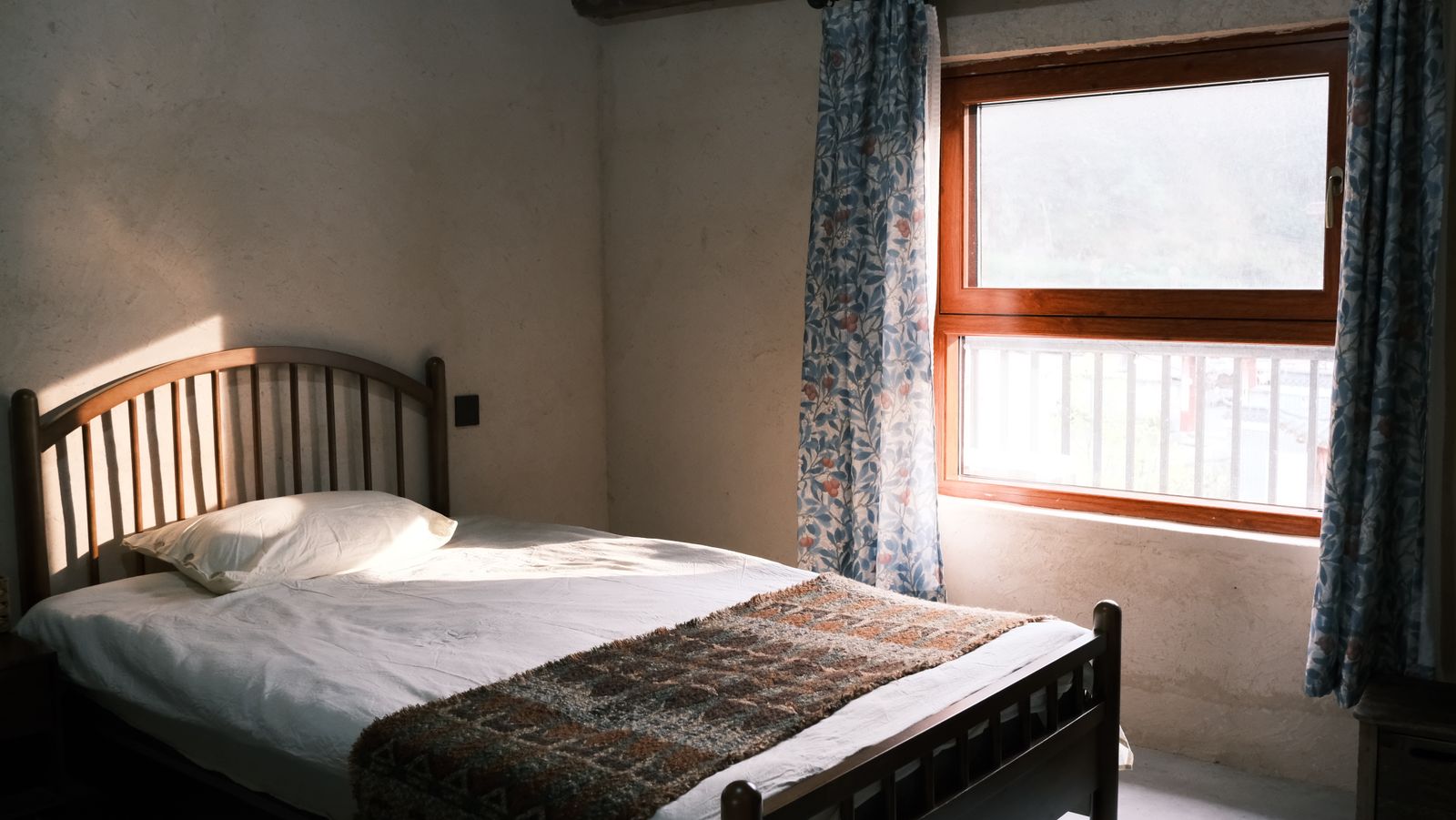 A wooden double bed by a tall window with floral curtains and morning sun