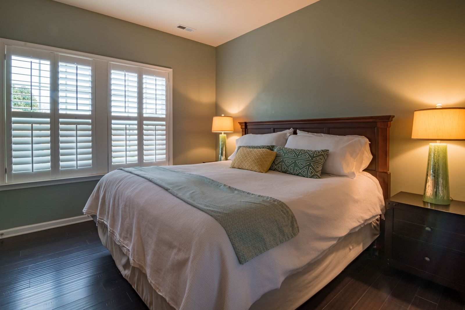 A double bed with a pale throw, two bedside lamps and white shutters