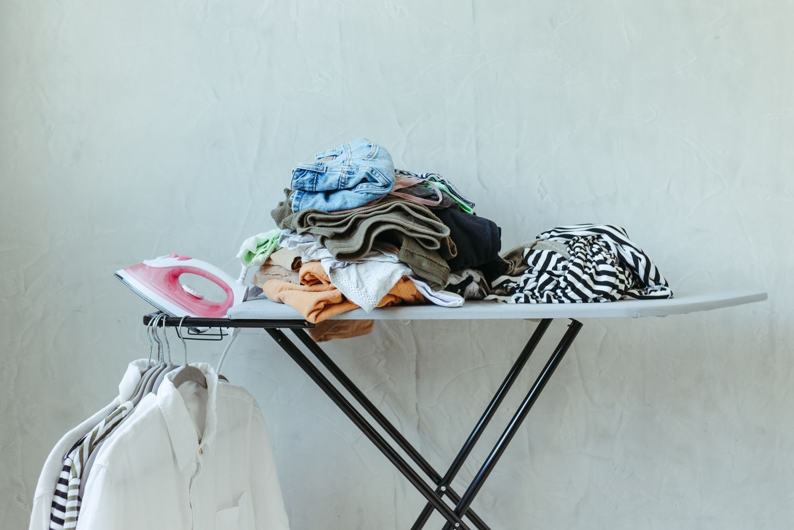 An ironing board with an iron, folded clothes and white shirts on hangers