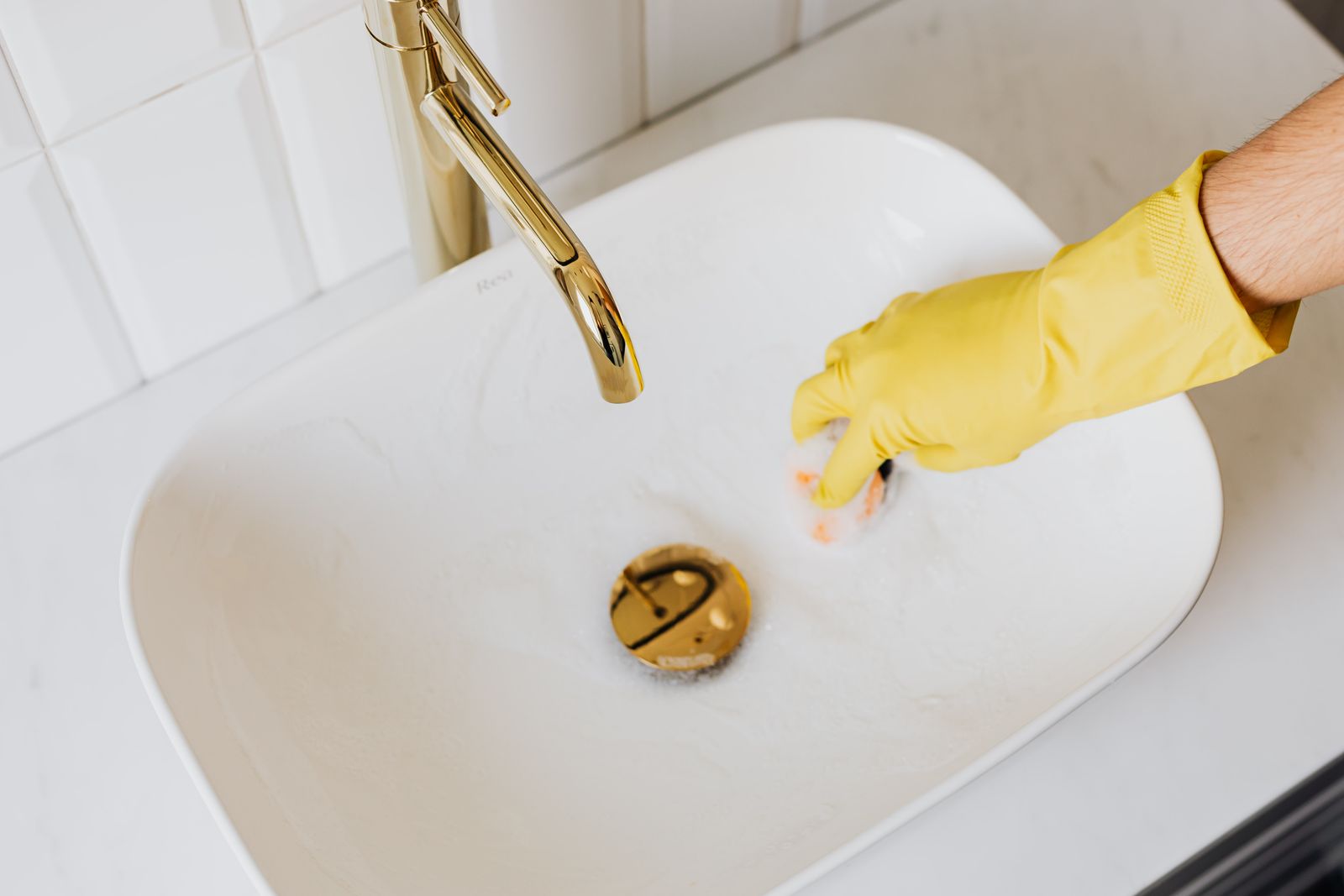A hand in a yellow rubber glove cleaning a white basin with a sponge