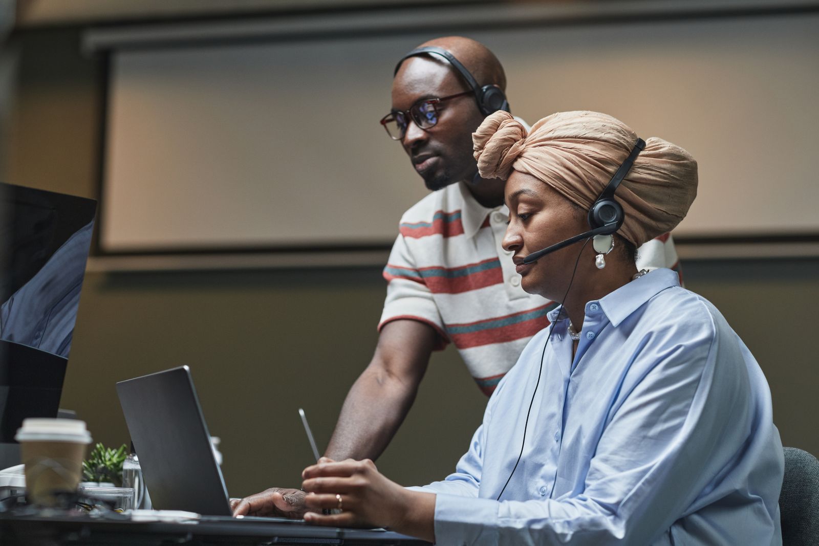 A support engineer with a headset helping a colleague at her laptop