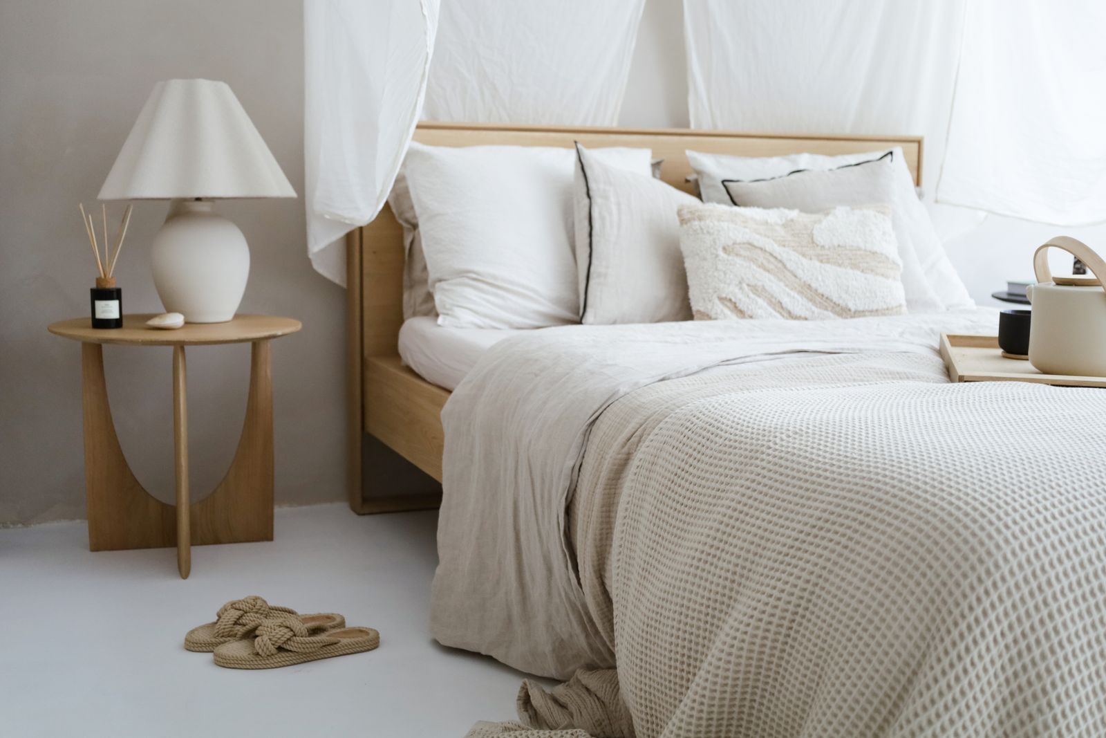 A bedroom with a wooden bed, white linen and a knitted throw, a round side table and a lamp