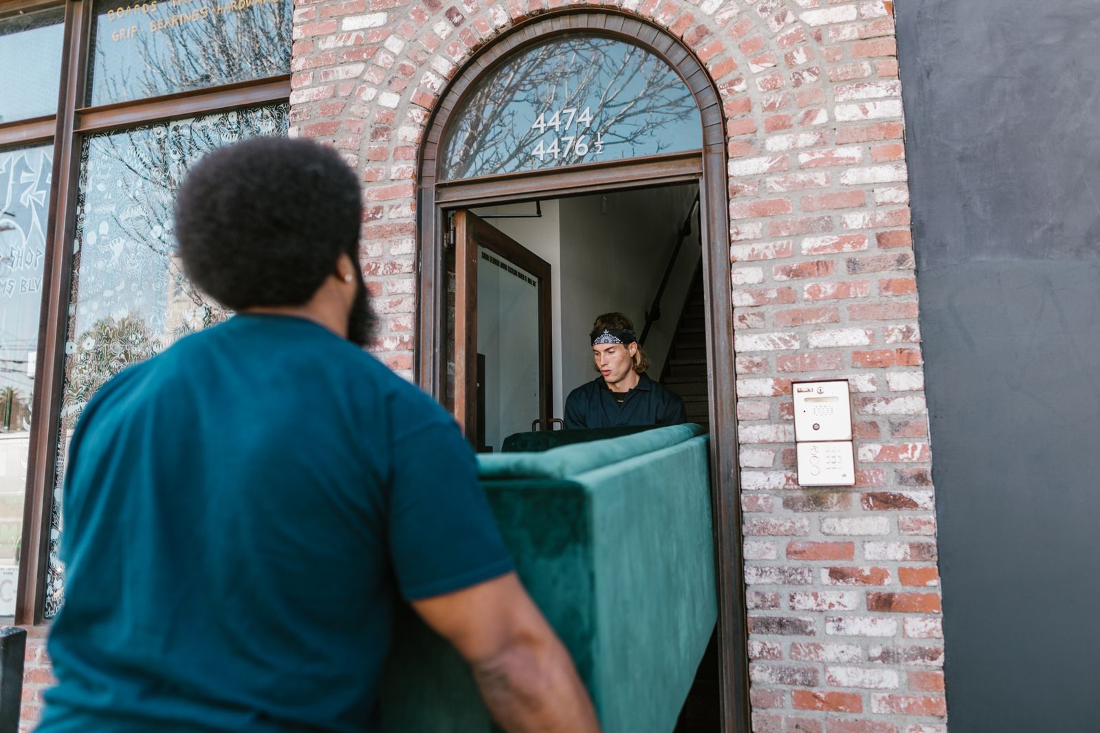 Two delivery men carrying a green velvet sofa through the arched brick doorway of a house