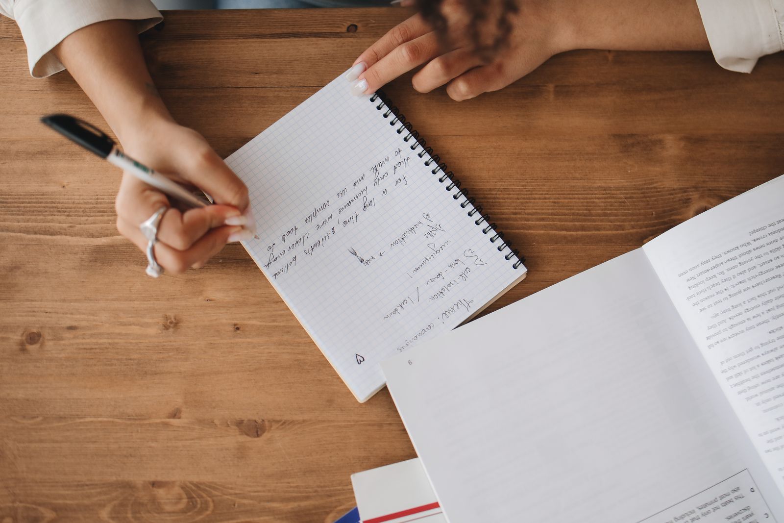 Hands taking notes in a notebook beside an open book on a wooden desk